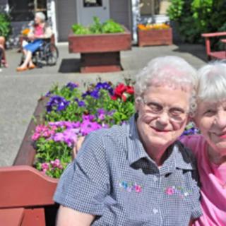 Two elderly women sitting together on a bench in a garden, enjoying a sunny day.