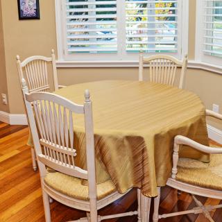 A cozy dining area with a round table covered by a beige tablecloth, surrounded by four chairs, and natural light streaming through the window blinds.
