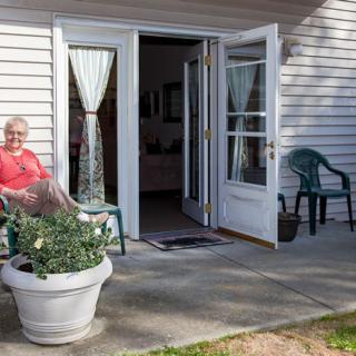 A person sitting on a porch chair outside a house with open doors and potted plants.