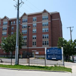 A multi-story brick residential building with a sign out front, surrounded by trees and a fence.