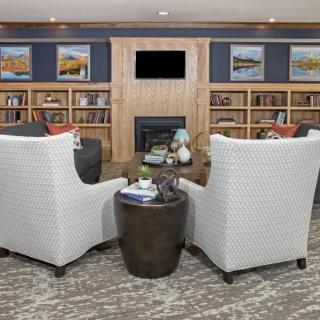 A cozy library setting with armchairs facing a fireplace, surrounded by bookshelves and artwork.