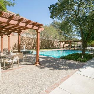 A serene outdoor pool area with pergolas and seating, surrounded by trees on a sunny day.