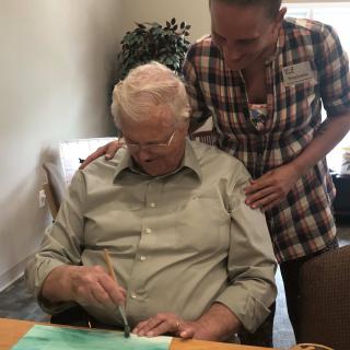 An elderly man engaged in a painting activity with the assistance of a caregiver.