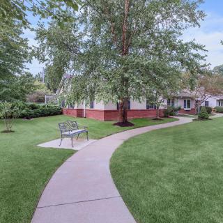 A curved pathway leads through a well-maintained lawn with trees and a bench, towards a house in the background.