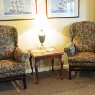 A cozy living room scene with two armchairs flanking a small wooden table topped by a lamp, and framed artwork on the wall.