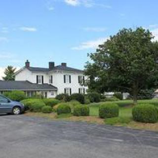 A large white house with a tree and bushes in the foreground, next to a parked car on a driveway under a blue sky.