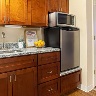 A small kitchen area with wooden cabinets, a countertop with a sink, a bowl of fruit, a refrigerator, and a microwave.