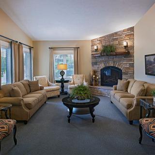 A cozy living room featuring plush sofas, a central coffee table, and a stone fireplace with natural light streaming in through large windows.
