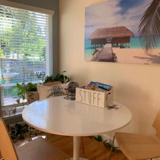 A cozy kitchen corner with a round table, surrounded by chairs and decorated with plants under warm sunlight. A tropical beach-themed wall art enhances the ambiance.
