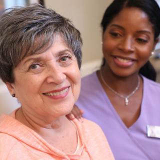 An elderly woman smiling warmly with a caregiver by her side.