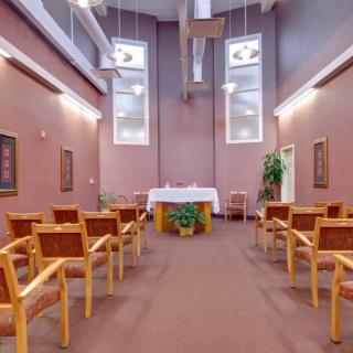 A serene chapel interior with rows of chairs facing an altar, illuminated by natural light from the large windows above.
