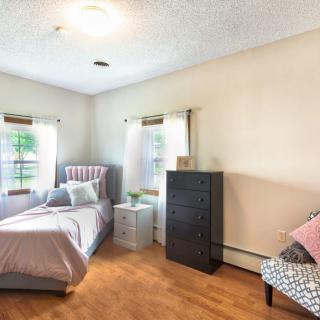 A cozy bedroom with a bed, dresser, and chair, featuring hardwood floors and natural light from two windows.