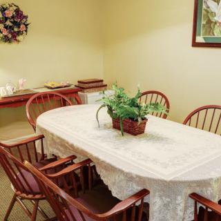A cozy dining room with a table covered by a lace tablecloth, surrounded by wooden chairs and adorned with a potted plant. The room features subtle decorations, including a wall painting and festive wreath.