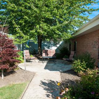 A peaceful garden area with a walkway, surrounded by lush trees and bushes, and featuring chairs on a patio.
