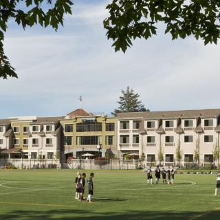 Students gather on a soccer field in front of a school building, under a cloudy sky.