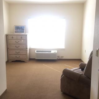 A simple, carpeted room with a dresser, armchair, and a large window letting in natural light.