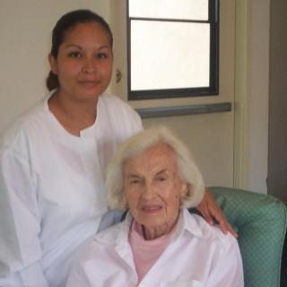 A nurse stands beside an elderly woman, both smiling, in a healthcare setting.