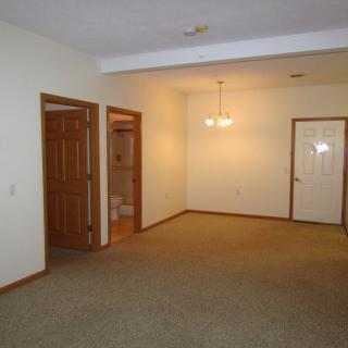 A vacant living space with carpet flooring and ceiling light fixtures, featuring doorways leading to other rooms.