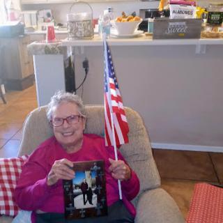 An elderly woman proudly holding a photo and an American flag, celebrating in her cozy home.