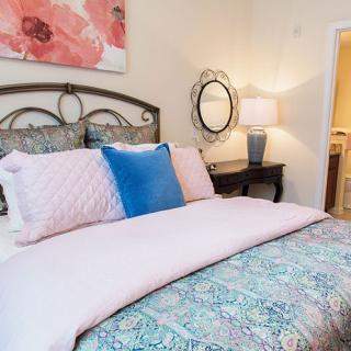 A cozy bedroom with a neatly made bed featuring pink and blue pillows, a bedside table with a lamp, and wall decor including a mirror and floral artwork.