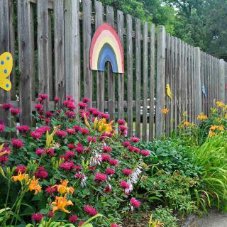 A vibrant garden with various flowers blooming alongside a wooden fence adorned with a rainbow and butterfly decorations.