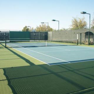 An outdoor tennis court with a net, surrounded by fences and equipped with lighting for games.
