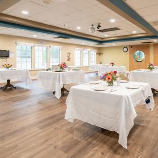A well-lit dining room or event space with neatly arranged tables covered in white tablecloths and decorated with flowers.
