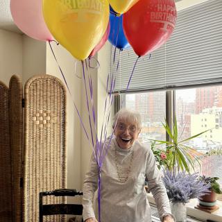 An elderly woman joyfully holding colorful balloons indoors, surrounded by plants and sunlight.