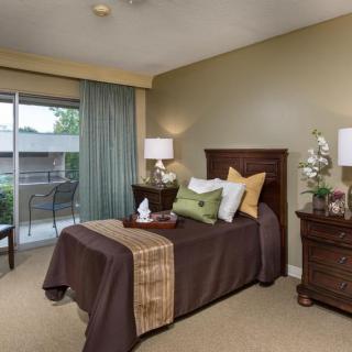 A cozy bedroom with a neatly made single bed, surrounded by two nightstands and lamps, featuring decorative pillows and a view of the balcony through large curtains.