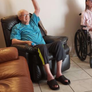 An elderly man in an armchair raises his hand, with a wheelchair visible nearby.