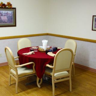 A small dining area with a round table covered in a red tablecloth and four chairs, decorated with a centerpiece, set against a wooden floor and framed paintings on the wall.