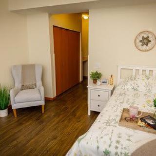 A cozy bedroom with wooden flooring, featuring a comfortable bed, a bedside table with decor, and an inviting armchair.