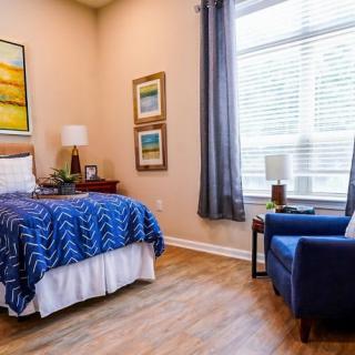 A cozy bedroom with a blue armchair, a neatly made bed, and large window with curtains.