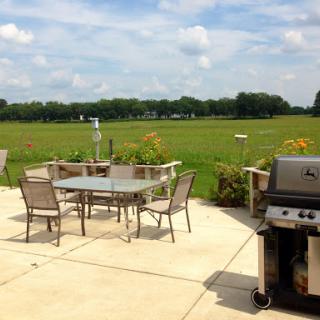 A serene outdoor patio set up with a grill, table, and chairs, surrounded by lush green fields under a bright blue sky.