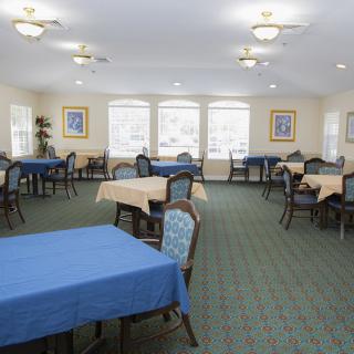 A spacious dining room with multiple tables and chairs, featuring large windows and a carpeted floor.