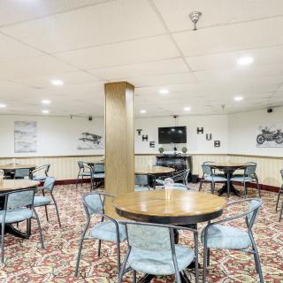 A spacious cafeteria with tables and chairs arranged on a carpeted floor, featuring wall decorations and a television.