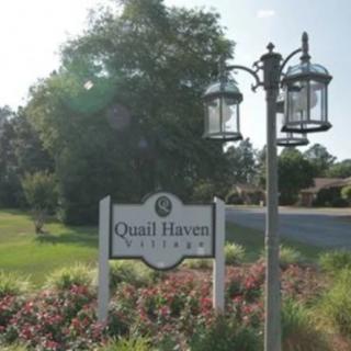 A sign for Quail Haven Village stands alongside a lamppost in a picturesque neighborhood setting.