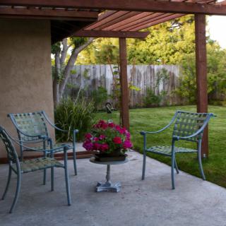 A cozy patio with metal chairs and a small table under a pergola, overlooking a lush garden.