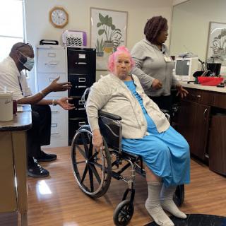 An elderly woman in a wheelchair being assisted by caregivers in a healthcare setting.