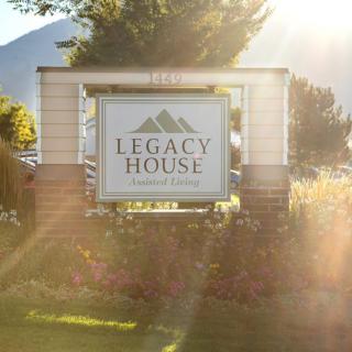 The Legacy House Assisted Living sign stands illuminated by sunlight in a scenic outdoor setting.