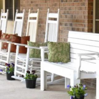 A row of white rocking chairs with cushions on a porch, accompanied by potted plants.