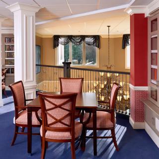 A cozy dining area with wooden furniture and blue carpeting, featuring large windows and decorative columns.