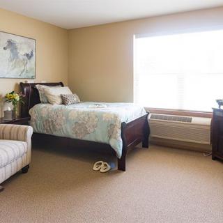 A cozy bedroom with a neatly made bed, an armchair, and a large window letting in natural light.