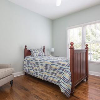 A cozy bedroom with a wooden single bed and patterned bedding, featuring a large window and an armchair on hardwood floors.