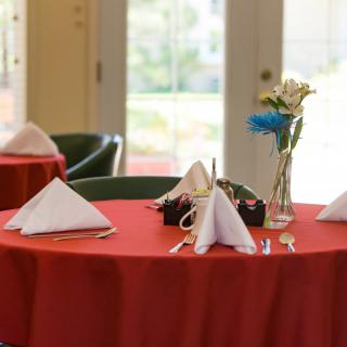 A restaurant table set with a red tablecloth, neatly folded white napkins, and a vase of flowers.