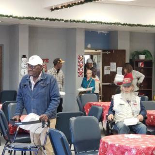 A group of people in a community setting, engaging in an indoor gathering with tables and chairs arranged around them.