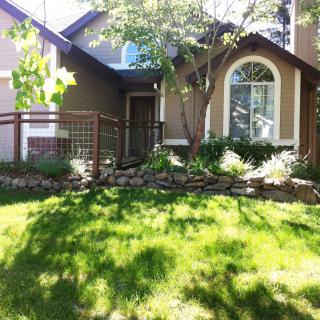 A charming house with a lush green lawn and garden, featuring large windows and shaded by trees.