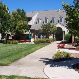 A sunny day view of a residential building with a well-maintained lawn and pathway surrounded by trees.