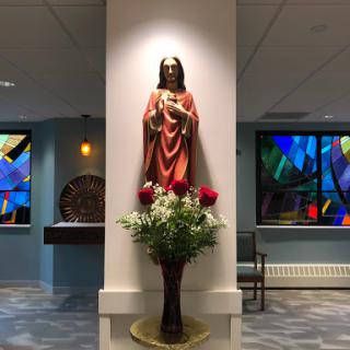 A serene interior space featuring a statue adorned with flowers, complemented by vibrant stained glass windows.