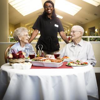 An elderly couple enjoys a meal at a restaurant, served by a smiling staff member.